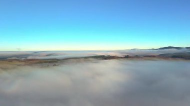Açık mavi gökyüzü altındaki bir sis denizinin havadan görünüşü, alçak bulutlar tepeler ve ormanlar üzerinden akıyor.
