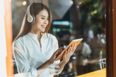 Çalışmak ve müzik dinlemek için pencere kenarındaki bir kafede elinde teknoloji tableti tutan ve kullanan genç Asyalı kadın portresi, internete bağlanarak çalışan serbest yazar ve girişimci.