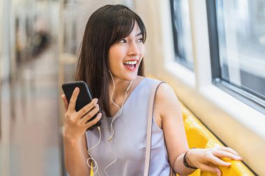 Genç Asyalı kadın yolcu metroda, Japonca, Çince, Kore yaşam tarzı ve günlük yaşam, banliyö ve ulaşım kavramında seyahat ederken akıllı cep telefonuyla müzik dinliyor.