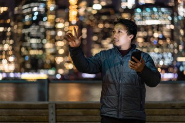 Paltolu bir Asyalı, akıllı cep telefonu kullanıyor. New York 'un bulanık fotoğrafı üzerinde gülücükler saçıyor. New York City' nin doğu nehri kıyısında.