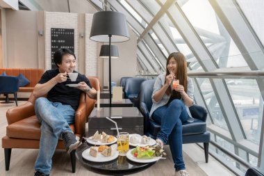 Asyalı çift, modern uluslararası havaalanı, seyahat ve ulaşım konseptinde uçak beklerken otellerde oturup yemek yiyor.