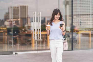 Asyalı bir kadın şehirde yürüyor, cep telefonunu kullanıyor ve elinde bir fincan kağıt kahve tutuyor. Alışveriş merkezi, yaşam tarzı ve eğlence, online alışveriş ve nakit ödeme konsepti