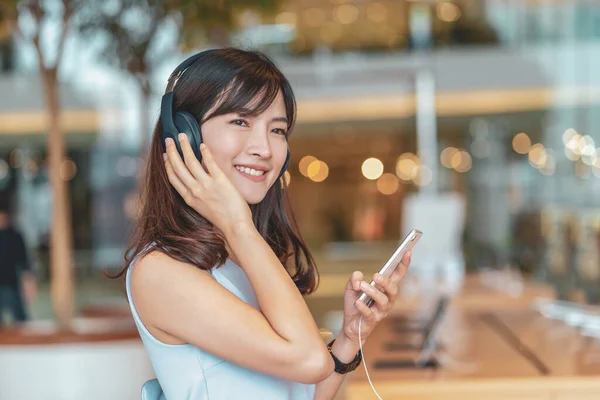 Asian woman using smart mobile phone and listening the music via Head cover headphones in department store or shop store, entertainment and lifestyle concept