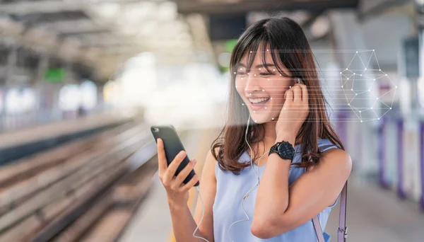 スマートフォンを介して顔認識を使用し 鉄道駅プラットフォームで音楽を聞いてアジアの女性 生体認証と人工知能の概念 ロイヤリティフリーのストック写真