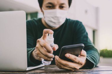 Bilgisayarla çalışmadan önce cep telefonunu temizlemek için dezenfektan alkol kullanan, Covid-19 salgını, sosyal mesafe ve sorumluluk kavramı için evden çalışan Asyalı adam.