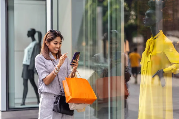 Çevrimiçi alışveriş siparişini kontrol etmek için akıllı cep telefonunu kullanan Asyalı kadın, mağazadaki cam mağazanın yanındaki kıyafetlerle birlikte mağazada mutluluk verici bir şekilde tamamlanıyor.