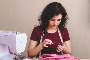 Mutlu ve çekici genç bayan terzi stüdyoda dikiş makinesine oturup dikiş dikiyor.