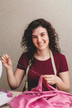 Mutlu ve çekici genç bayan terzi stüdyoda dikiş makinesine oturup dikiş dikiyor.