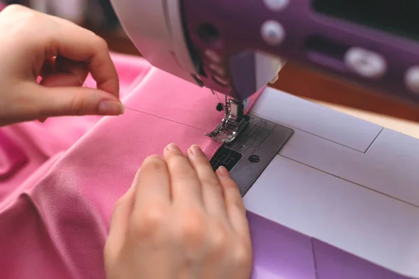 Happy attractive young woman seamstress sitting and sews on sewing machine on light background. Concept of sewing, handmade goods. Advertising of studio.