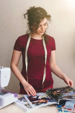 Mutlu ve çekici genç bayan terzi stüdyoda dikiş makinesine oturup dikiş dikiyor.