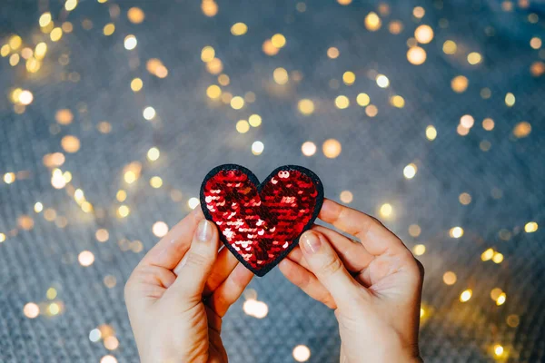 Lantejoulas vermelhas coração em mulheres mãos no fundo cinza com luzes bokeh. — Fotografia de Stock