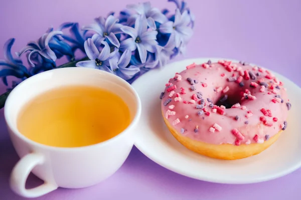 Pink sugar glazed donut, cup of tea and purple hyacinth flower on colorful background. — Stock Photo, Image