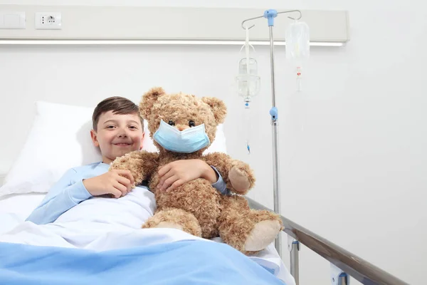 smiling child in hospital bed with teddy bear wearing protective mask, corona virus covid 19 protection concept,