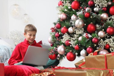 Mutlu noeller ve mutlu tatiller, çocuk evde bilgisayarla aydınlanmış ve süslenmiş ağacın ve paketlenmiş hediye paketlerinin yanında kal.