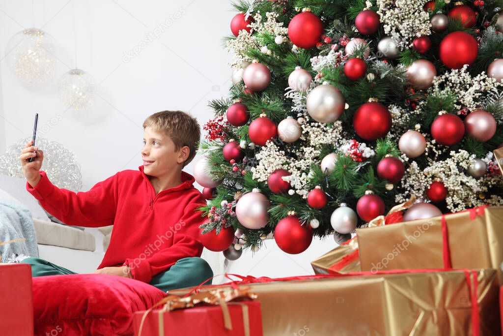merry christmas and happy holidays, child stay at home with mobile phone near the illuminated and decorated tree and wrapped gift packages