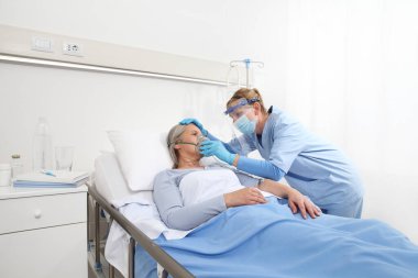 Hemşire, hastane yatağında yatan yaşlı bir kadına koruyucu eldivenler ve maske takarak oksijen maskesi takıyor. Coronavirus Covid 19 koruma kavramı.