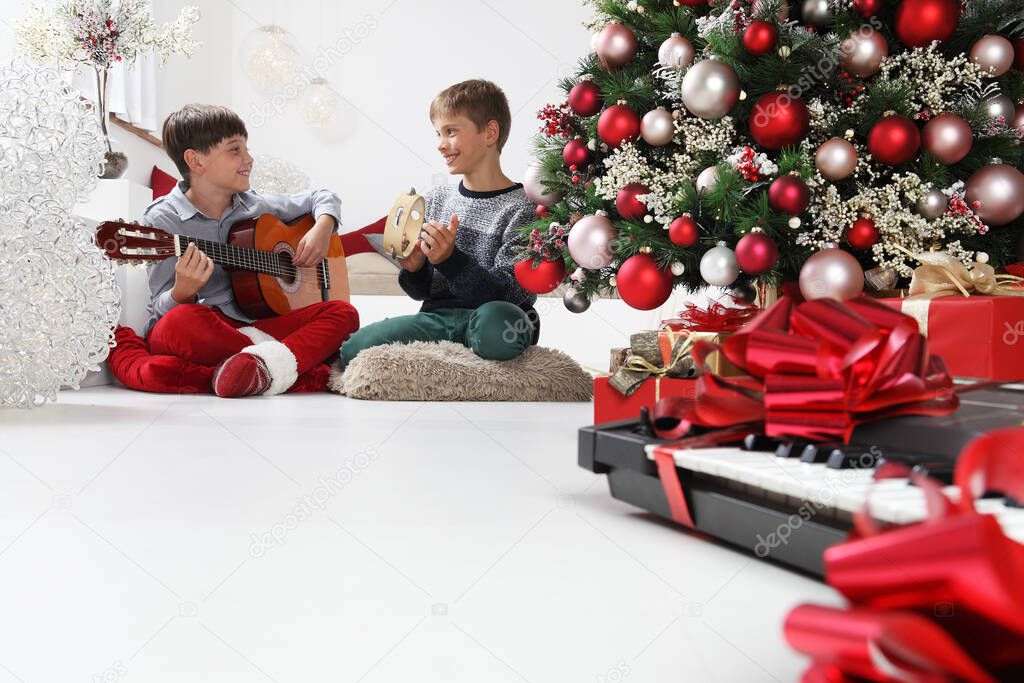 merry christmas and happy holidays, children play guitar and tambourine near the christmas tree with wrapped gift packages and musical instruments, at home in the living room sitting on the floor