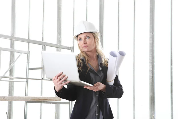 woman architect or construction engineer with computer laptop wear helmet and holds blueprint inside a building site with windows and scaffolding in the background