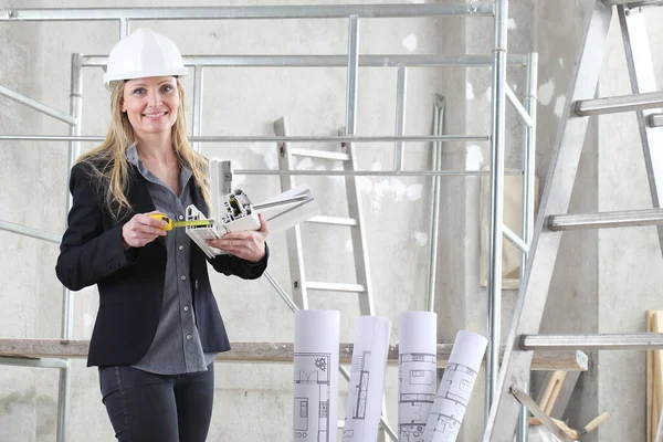 smiling woman architect or construction interior designer with with meter measuring white windows cutaway profile inside a building site with ladder and scaffolding in the background