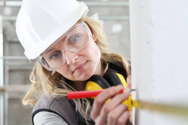 woman construction worker work with meter tape and pencil, measure wall in interior building site, wearing helmet, glasses and hearing protection headphones