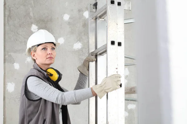 建筑工人妇女头像 头戴白色头盔 戴听力保护耳机 手持梯子 脚手架在建筑的内部背景 — 图库照片