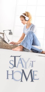 Bilgisayarlı ve kulaklıklı genç bir kadın, dizüstü bilgisayar monitörüne bak, beyaz zemin üzerinde izole edilmiş evde mesajla kal, Coronavirus covid-19 önleme için sosyal medya kampanyasında kal