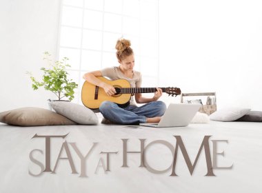 Bilgisayarlı genç bir kadın gitar çalıyor, dizüstü bilgisayar ekranına bakıyor, beyaz zemin üzerinde izole edilmiş evde mesajla kalıyor, Coronavirus covid-19 korunması için sosyal medya kampanyasında kalıyor.