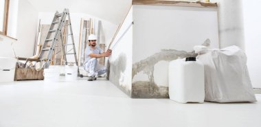 İnşaat işçisi sıva adam, inşaat alanında aletlerle ve zeminde inşaat malzemeleriyle birlikte mezura ile ölçü bandı ile duvarı ölçüyor.