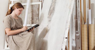 Tuvalin önünde oturan ve parlak beyaz stüdyodaki eskiz defterlerine bohem şıklığında resim çizen genç bir kadın ressam.