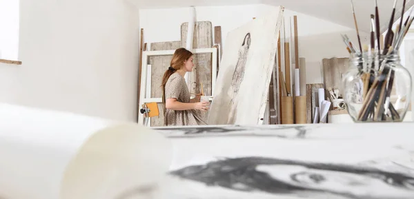 年轻女画家坐在画布前 穿着波希米亚式时髦服装坐在明亮的白色画室里作画的室内照片 其前景以背景为重点 — 图库照片