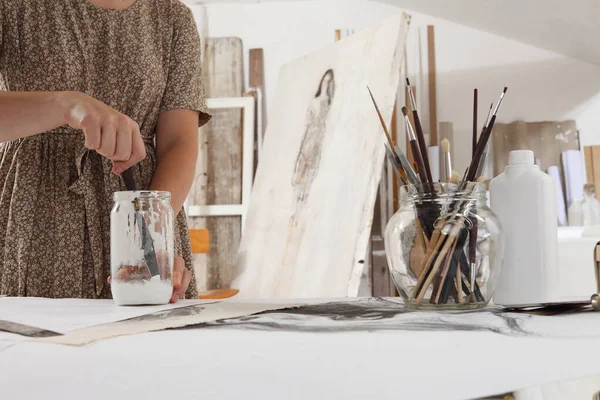 Indoor shot of half length cropped body and hands with no head of female artist in bright white studio while cleaning brushes wearing bohemian chic clothing