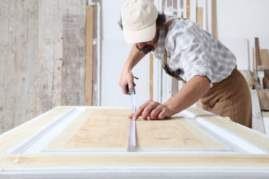 Marangozluk atölyesinde ahşap işi yapan erkek marangoz, ahşap kapıyı mezurayla ölçüyor.