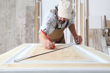 Marangozluk atölyesinde ahşap işçiliği yapan erkek marangoz, ahşap kapı üzerinde ölçü bandı ve kurşun kalemle tedbirler alıyor.