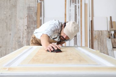 Marangozluk atölyesinde çalışan erkek marangoz zımpara kağıdıyla ahşap bir kapıyı zımparalıyor.