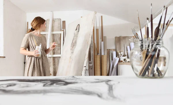 年轻女画家坐在画布前 穿着波希米亚风格服装在明亮的白色画室里作画的室内照片 — 图库照片