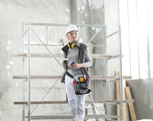 woman worker builder to coffee break with mobile phone, wearing helmet, glasses, bag tools and ear protection headphones, scaffolding construction site indoors on background