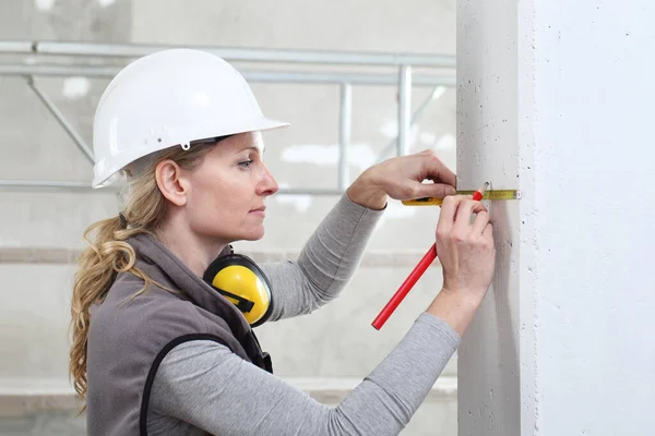 woman construction worker work with meter tape and pencil, measure wall in interior building site, wearing helmet and hearing protection headphones