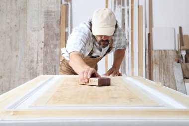 Male carpenter working the wood in carpentry workshop, sanding a wooden door with sandpaper, wearing overall and cap clipart