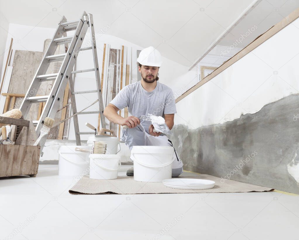 man plasterer construction worker at work, takes plaster from bucket and puts it on trowel to plaster the wall, wears helmet inside the building site of a house