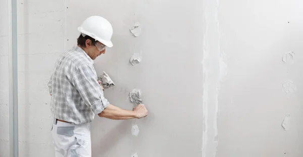 man drywall worker or plasterer putting plaster on plasterboard wall using a trowel and a spatula, fill the screw holes, wearing white hardhat and safety glasses.