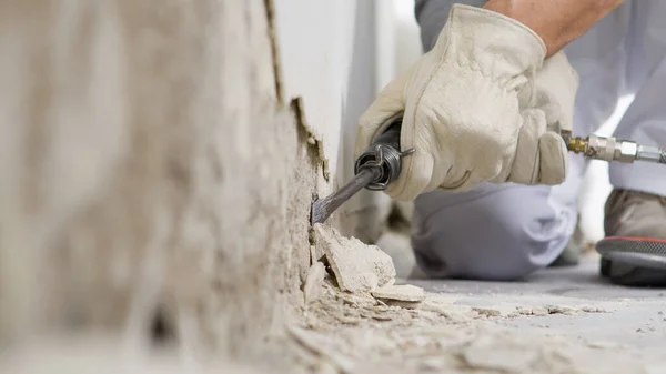 house renovation concept, construction worker breaks the old plaster of the wall with pneumatic air hammer chisel, close up with rubble on floor