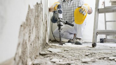 Ev yenileme konsepti. İş yerinde koruyucu eldivenleri ve sarı miğferi olan inşaat işçisi beyaz arka planda kaya matkabı, alçı moloz ve merdiven ile duvarı kırıyor..
