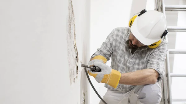 house renovation concept, construction worker breaks the old plaster of the wall with pneumatic air hammer chisel, wears gloves, helmet and safety yellow headphones , close up with white copy space
