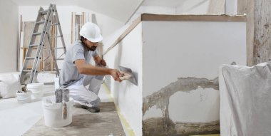 İşteki adam sıva işçisi, kovadan alçı alıyor ve duvara yapıştırmak için malaya yapıştırıyor, bir evin inşaat alanında kask takıyor.