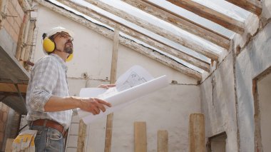 Mimar, miğfer ve kulaklık takıyor. Şablona bakıyor. İnşaat projesinin planını gözden geçiriyor. İnşaat alanının arka planında çatının eski ahşap kirişleri var.