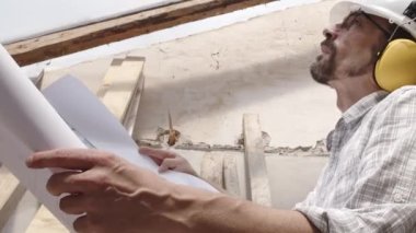 Mimar, miğfer ve kulaklık takıyor. Şablona bakıyor. İnşaat projesinin planını gözden geçiriyor. İnşaat alanının arka planında çatının eski ahşap kirişleri var.
