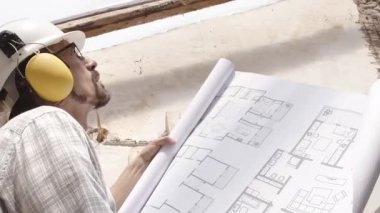 Mimar, miğfer ve kulaklık takıyor. Şablona bakıyor. İnşaat projesinin planını gözden geçiriyor. İnşaat alanının arka planında çatının eski ahşap kirişleri var.