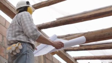 Mimar, miğfer ve kulaklık takıyor. Şablona bakıyor. İnşaat projesinin planını gözden geçiriyor. İnşaat alanının arka planında çatının eski ahşap kirişleri var.