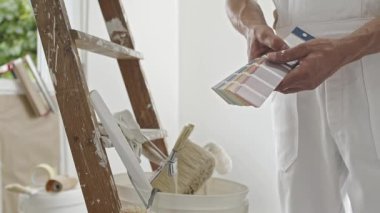 Ev boyacısı dekoratörünün elleri numune örneğini, yenilemek için evin çalışmasını, arka planda boya fırçası ve kova olan ahşap bir merdiveni kullanarak rengini seçer.