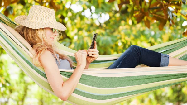 用智能手机对着金发碧眼的女人微笑 在花园里的吊床上放松地躺着 利用业余时间和暑假来上网或者用社交媒体和朋友聊天 — 图库照片
